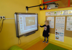 Dziewczynka stoi pod tablicą interaktywną i wskazuje ilustrację przedstawiającą kwiaty.